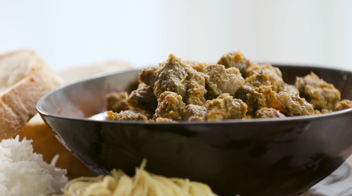 A bowl of meat curry with sides of rice, bread, and noodles.